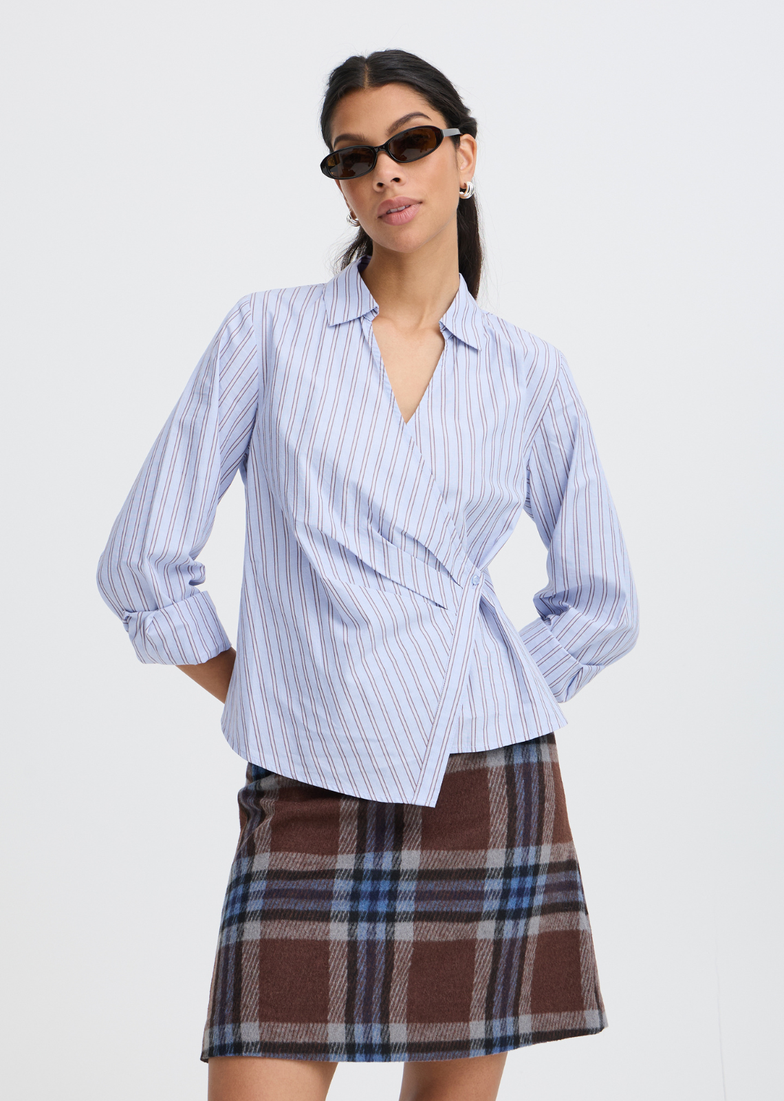 Reimagine classic shirting with this modern wrap blouse. Crafted in light blue with sophisticated vertical stripes, it features a wrap design that secures along the waist, highlighting your natural shape with dynamic crossover pleating and an asymmetrical hemline. The crisp collar and deep V-neck keep it clean and refined, making it the perfect statement piece put with trousers or pair with seasonal skirts.