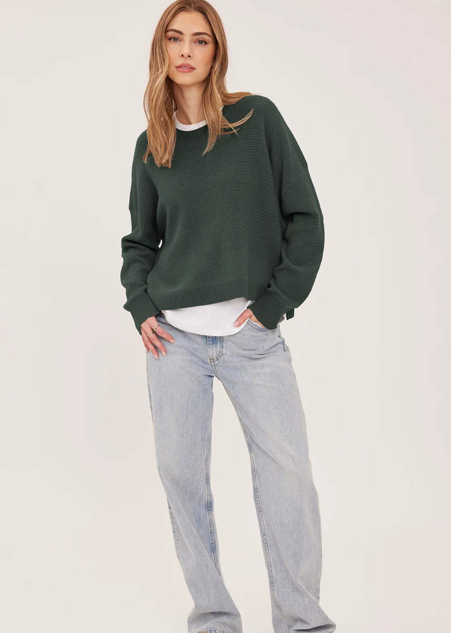 The Axel sweater is made of a soft yarn in a waffle knit pattern. Features include a crew neckline, exposed seam details, and side slits.