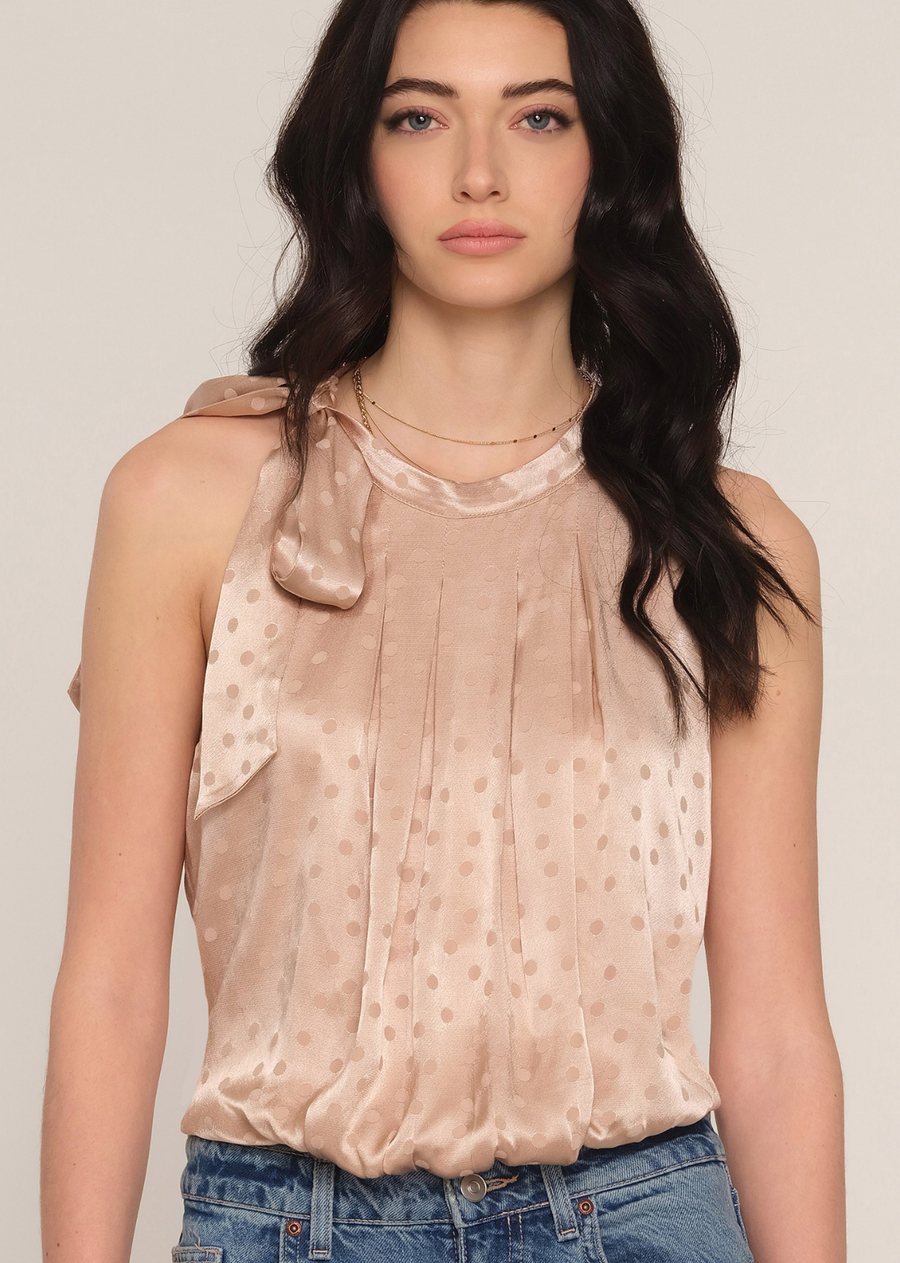 The Annicka Top is satin-finish sleeveless blouse in a champagne hue. The top is elevated by a feminine, oversized bow tie detail on the left shoulder and a high, gathered neckline with delicate vertical pleats creating a soft, draped bodice. The fabric has a subtle tonal polka dot texture. This blouson-style top features an elasticized hem, tucked into the jeans, highlighting the waist. The overall look blends refined elegance with casual cool.