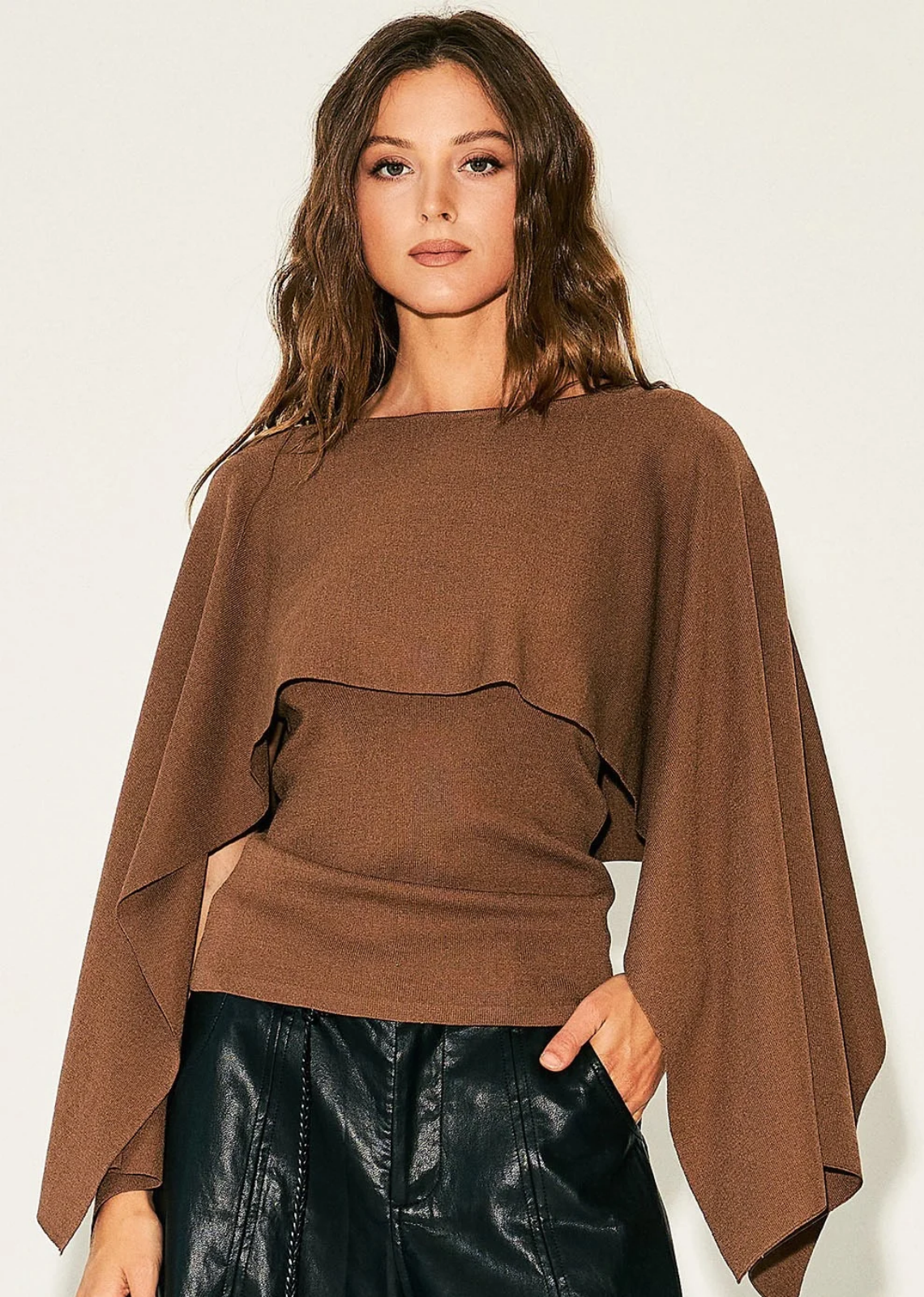 Elevate your transitional wardrobe with this cape overlay knit top. Designed with a chic layered construction, this piece features a fitted, sleeveless base paired with a dramatic, flowing caped overlay that drapes over the shoulders and arms for a striking silhouette. The soft, warm neutral tone makes it an exceptionally versatile piece to pair with everything from sleek faux-leather trousers to tailored skirts.
