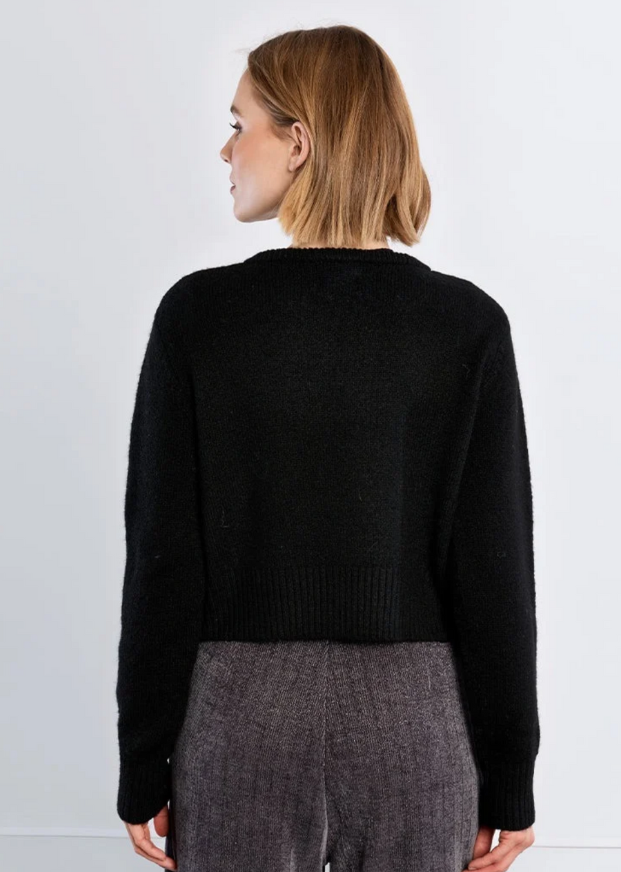 Effortlessly modern and sophisticated, this classic black knit cardigan is designed with a sleek open-front silhouette that captures modern elegance. Featuring a clean crew neckline secured by a delicate silver accent closure at the collar, it offers an eye-catching focal point that doubles as built-in jewelry. Crafted with a soft, cozy texture, ribbed trim, and a flattering slightly cropped length, this versatile piece transitions seamlessly from day to night. Pair it over a fitted printed layer and wide-l