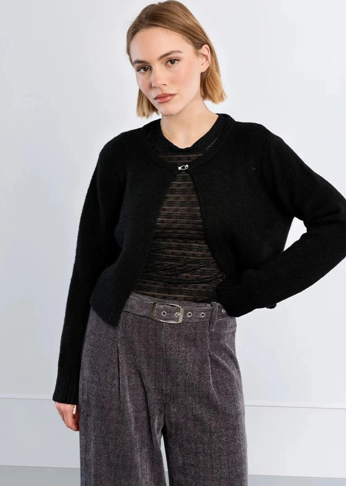 Effortlessly modern and sophisticated, this classic black knit cardigan is designed with a sleek open-front silhouette that captures modern elegance. Featuring a clean crew neckline secured by a delicate silver accent closure at the collar, it offers an eye-catching focal point that doubles as built-in jewelry. Crafted with a soft, cozy texture, ribbed trim, and a flattering slightly cropped length, this versatile piece transitions seamlessly from day to night. Pair it over a fitted printed layer and wide-l