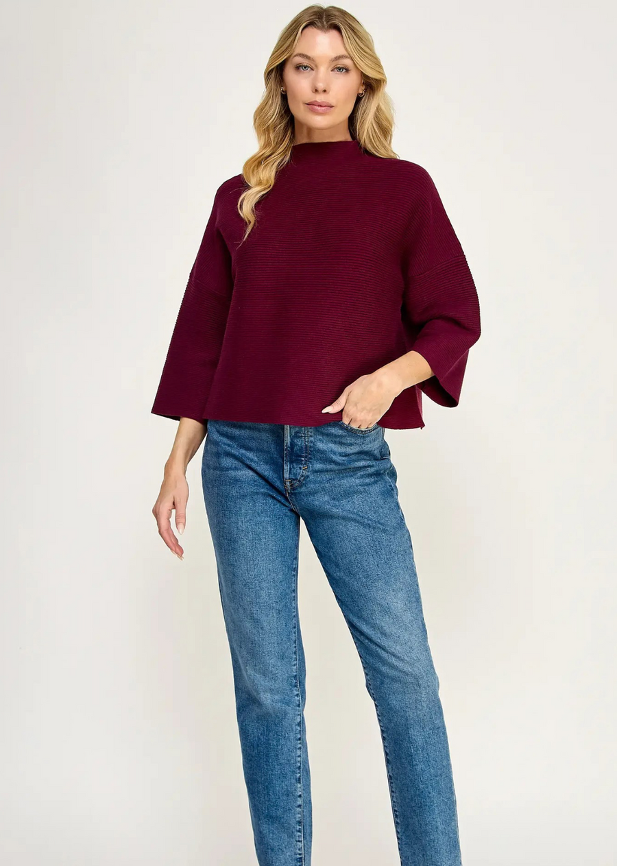 A modern mock-neck knit sweater in a rich forest green tone, featuring a clean horizontal rib texture and relaxed silhouette. Designed with dropped shoulders and modified three-quarter length sleeves, this piece offers a boxy, contemporary drape perfect for effortless layering.