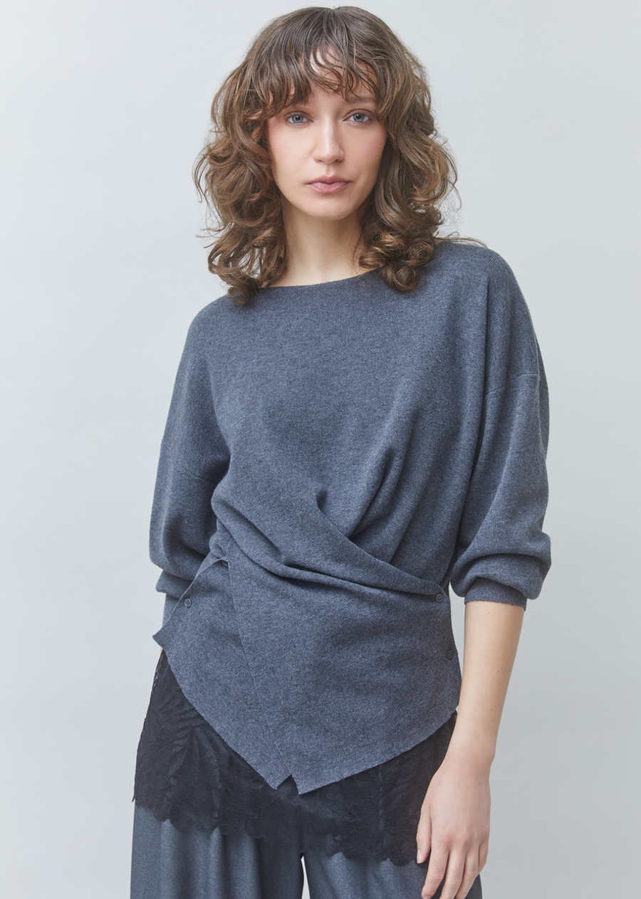 Elevate your cool-weather wardrobe with modern architectural tailoring. Crafted in a charcoal fine-gauge knit, this sweater features a soft boat neckline, drop shoulders, and full length relaxed sleeves with ribbed cuffs.

The standout feature is its artfully draped crossover front, creating an asymmetrical overlay complete with subtle side button detailing and a geometric point hem. Designed with a loose, flattering silhouette, it layers effortlessly over high-waisted trousers or lace-trimmed slip skirts f