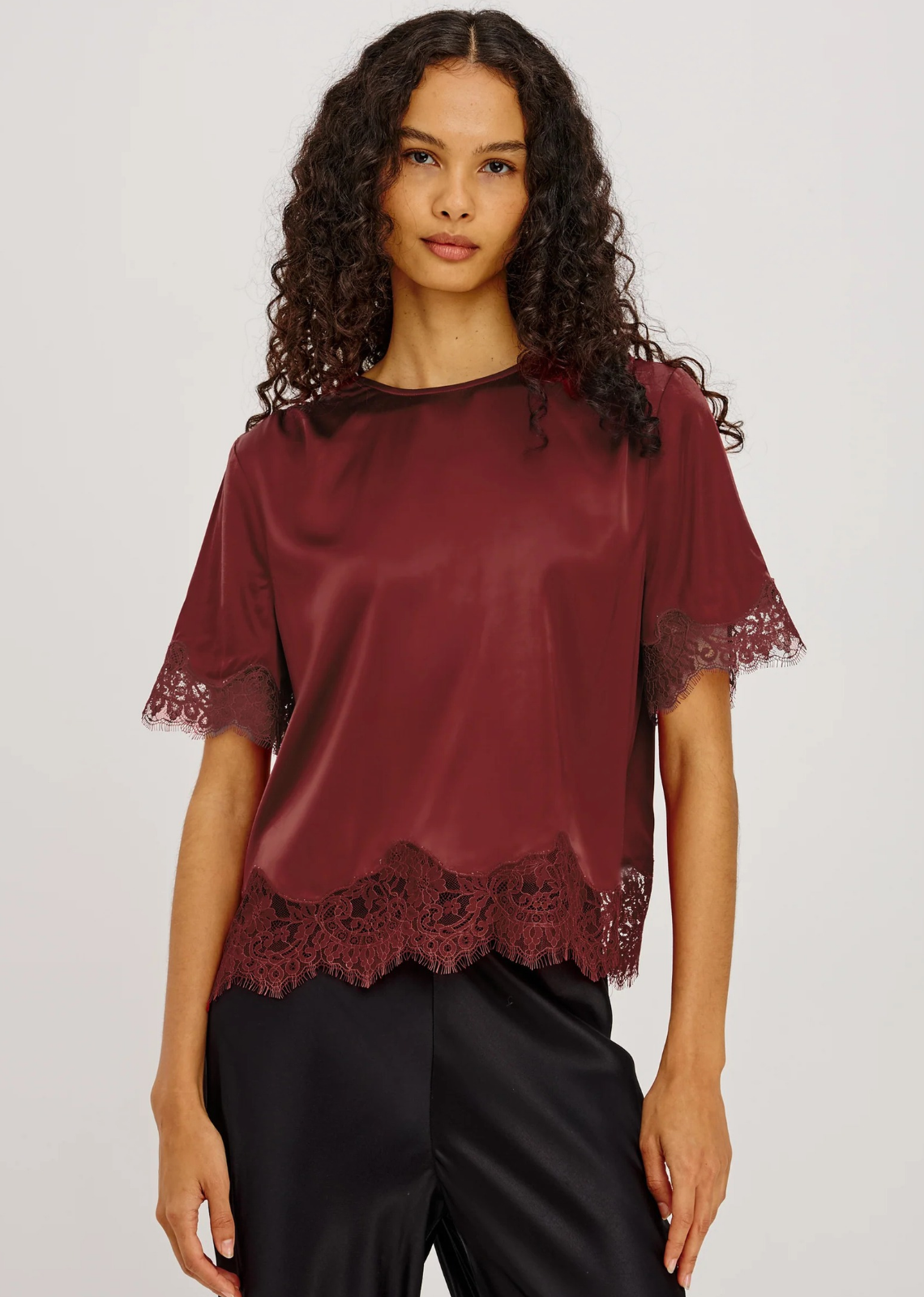 The Sonora Top is cut from a lightweight satin blend with a fluid drape and a loose, boxy fit. It features a crew neckline, shirred shoulders, short sleeves, a button-loop keyhole at the back neck, and tonal lace trim.