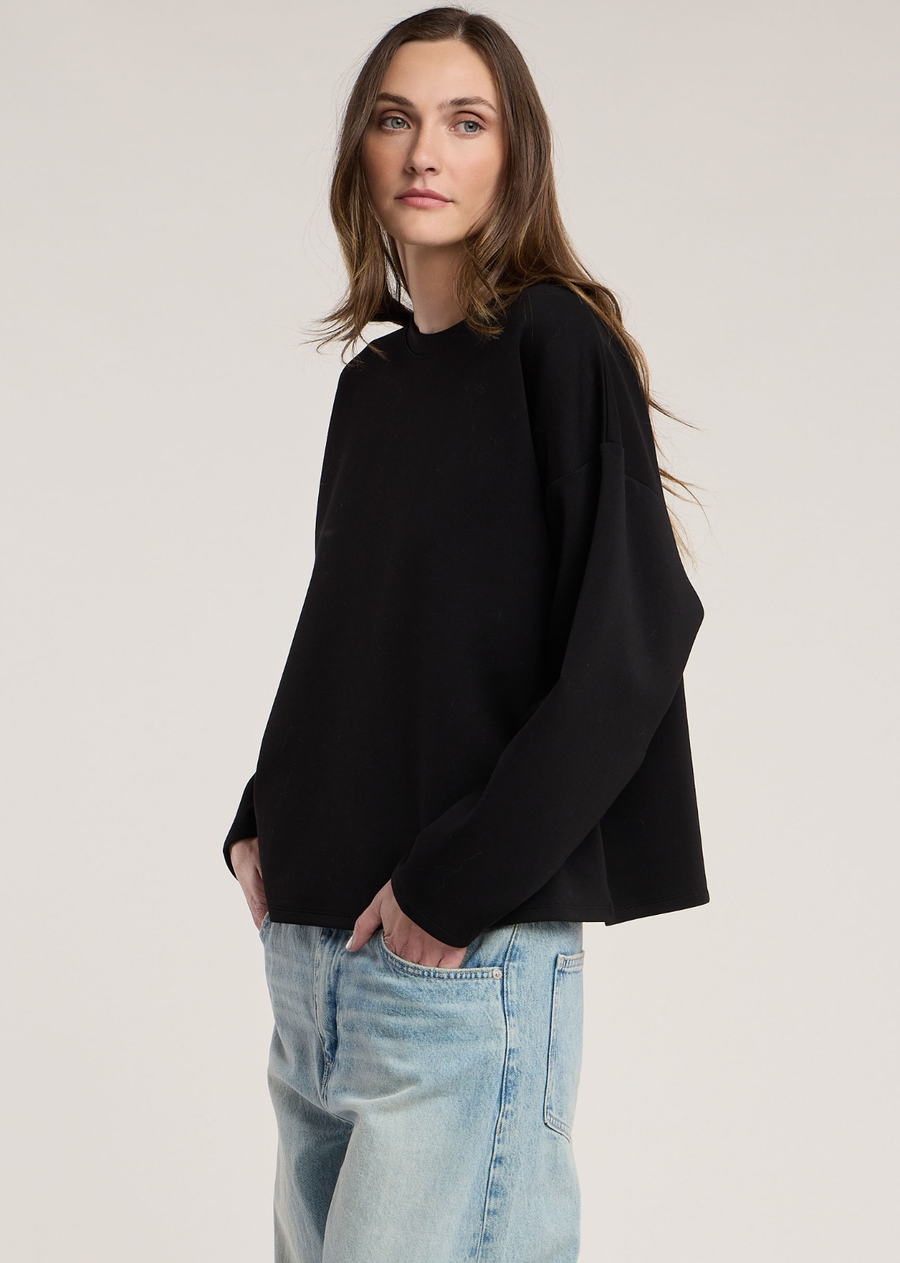 This long-sleeve black top offers a clean, architectural silhouette crafted from a smooth, structured scuba knit material. The fabric gives the garment a soft, spongey weight that holds its shape nicely while maintaining a comfortable, relaxed fit.
