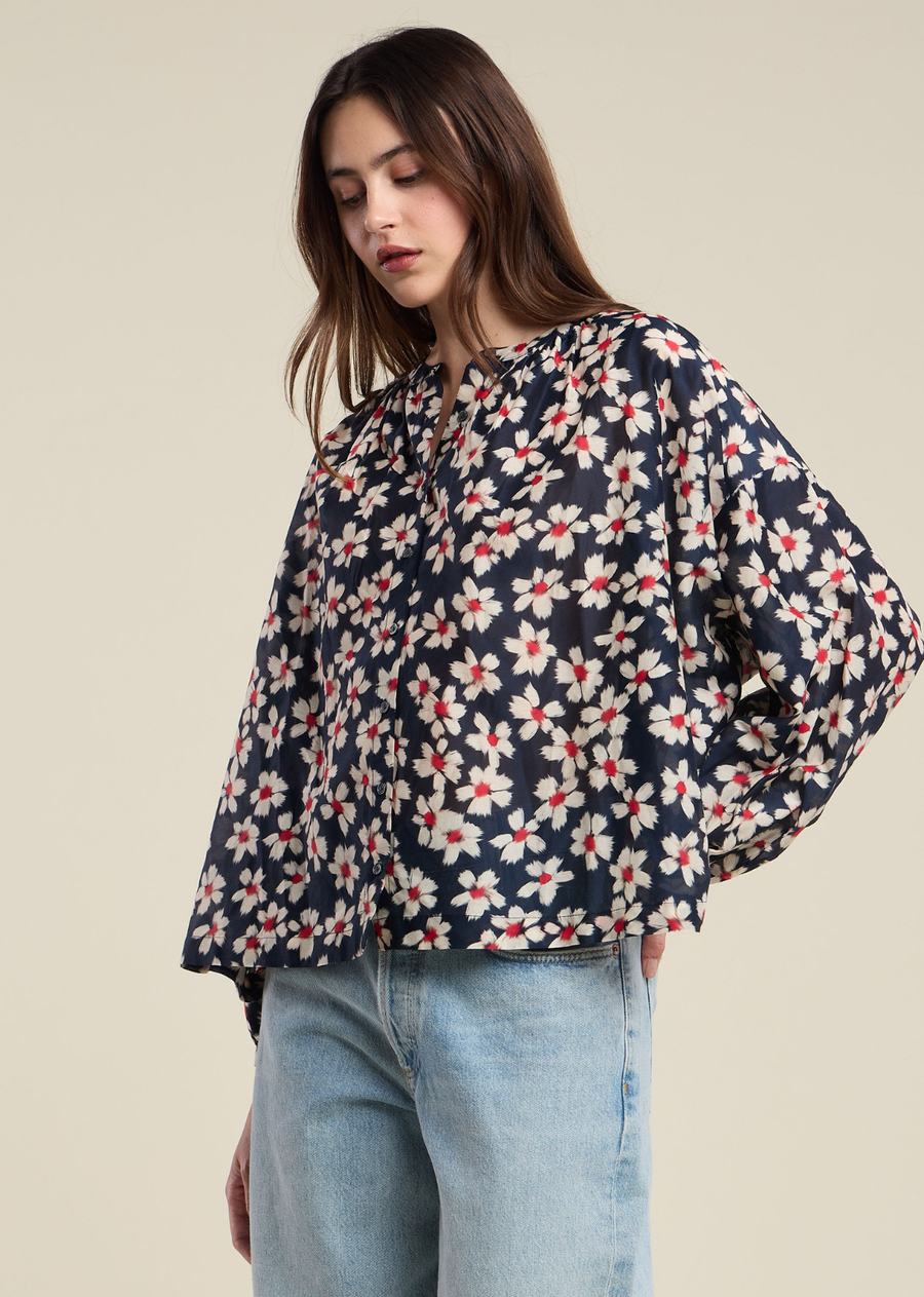 This fluid, lightweight top features a graphic floral pattern of cream and crimson red petals scattered across a dark navy backdrop. Designed with a split notch neckline and subtle gathering along the collar, it offers a romantic, airy silhouette that drapes into a relaxed, boxy cut. Voluminous long sleeves feature gathered cuffs to create a soft, puffed look, while a discreet button-down front maintains the continuous flow of the print.