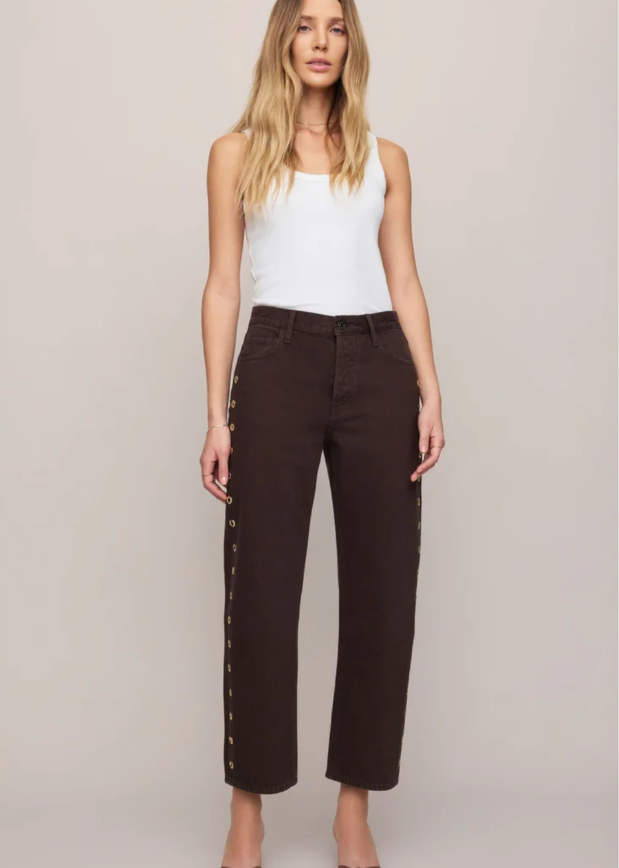 Rich brown denim, gold grommet details, and a relaxed bowed ankle shape give these jeans the kind of personality that makes the rest of the outfit feel easy. Crafted from rigid 100% cotton denim with subtle peekaboo openings along the sides, they feel playful, modern, and right at home in your closet for fall.