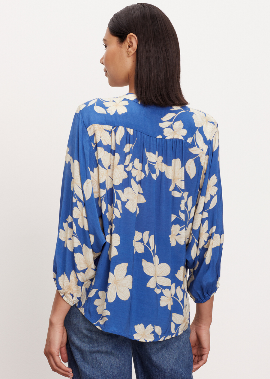Velvet Destina Top. This button-up blouse features delicately puffed sleeves and elastic cuffs for a refined touch. The daylily print adds a whimsical element, and the flattering scooped hemline enhances the silhouette. With a subtle hint of sheer, this blouse is the epitome of chic charm.