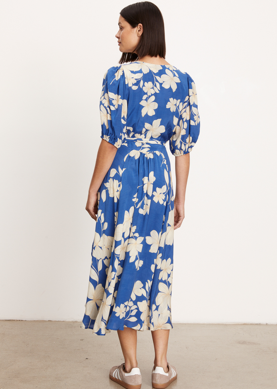 Velvet Kai Wrap Dress This dress features a flattering wrap dress style with a v-neckline and a midi-length, creating a timeless silhouette. The puff sleeves with elastic cuffs add a touch of elegance, while the daylily print enhances the overall charm.